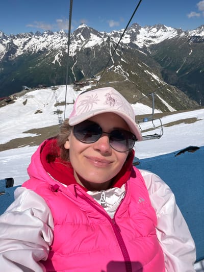 A medium shot selfie shows a woman wearing a pink puffer vest over a white jacket and a pink baseball cap, looking at the camera. She is wearing sunglasses. The background is a mountain landscape with snow-capped peaks and a ski lift. The location is a ski resort in Верхне-Тебердинское сельское поселение, Russia. The sky is clear blue with some scattered clouds. The overall scene is bright and sunny.