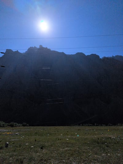 The image depicts a vast, dark mountain range silhouetted against a bright, hazy sky. The sun is a prominent, almost blinding white orb in the upper left quadrant, casting a strong glare. Several thin, dark power lines stretch horizontally across the frame, partially obscuring the mountain's features. These lines are strung between unseen poles, creating a sense of infrastructure within the natural landscape.
The mountain itself is largely devoid of detail due to the low light and shadow, appearing as a solid, dark mass. However, a series of lighter, horizontal stripes are faintly visible on its face, suggesting man-made terracing or possibly reflective elements catching the light. The terrain at the base of the mountains transitions into a grassy field, dotted with scattered rocks and debris. The grass is a mix of green and brown, indicating a dry or somewhat sparse growth. In the distance, beyond the field, a line of trees or shrubs is visible, forming a dark, irregular border. The overall atmosphere is one of a stark, natural landscape with subtle human intervention. The location is identified as Matayeen, India.