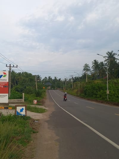 The image depicts a paved road in the city of Payabeunyot, Indonesia. On the left side of the road, a sign for Pertamina, a gas station, is visible. The sign displays prices for different fuel types, including Pertamax with a price of 15.950 for RON 92. Beyond the gas station, lush green vegetation and palm trees line the roadside, characteristic of the tropical environment. The road itself is a two-lane asphalt highway with a white line marking the center. Several individuals on motorcycles are seen traveling along the road. The sky is overcast with a cloudy expanse, suggesting a diffused light condition. The overall scene conveys a sense of a typical day on a rural Indonesian highway, characterized by local businesses, transportation, and abundant natural greenery.