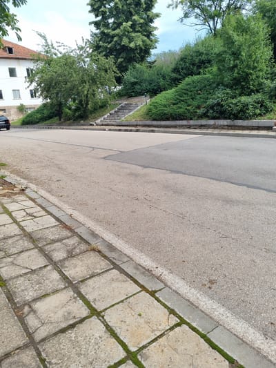 The image captures a street scene in Belogradchik, Bulgaria, with a focus on the road, sidewalk, and a vegetated slope. In the background, a white building with multiple windows and a reddish-brown tiled roof is visible on the left. Adjacent to the building, a tree with dense green foliage stands. Further to the right, a paved staircase leads up a steep, tree-covered slope. The road in the foreground is asphalt, with a white line marking its edge. To the left of the road is a worn concrete sidewalk composed of square slabs, with grass and moss growing in the cracks. A black car is parked on the left side of the road. The overall environment appears to be an urban setting with abundant greenery.