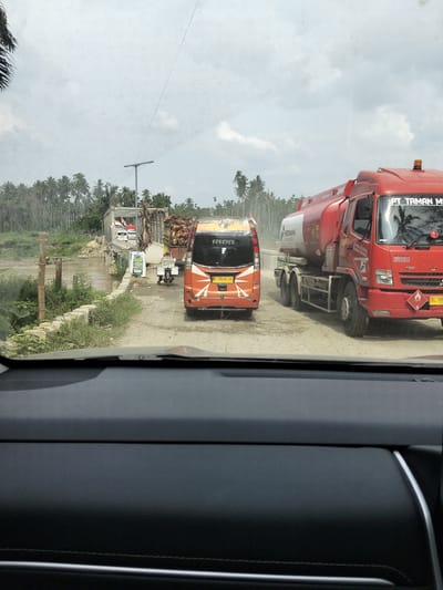 The image captures a traffic scene in Teupinreudeup, Indonesia, on a cloudy day. The foreground shows the interior of a car, suggesting the photo was taken from the driver's or passenger's seat.
The main subjects are vehicles on a dirt road. In the center, an orange minivan with the word "FOREVER" visible on its rear is stopped. To its right, a large red tanker truck with "PT TAMAN M" and "PERTAMINA" written on it is also present. The tanker truck has hazard symbols, including one with the number "3". Further back, a truck loaded with what appears to be palm tree fronds is visible near a bridge. Other vehicles and a lamppost are also present on the road.
The setting is a rural area with lush vegetation, including many palm trees, bordering the road and river. A bridge crosses the water, and the overall impression is that of a somewhat remote location. The weather appears overcast, with a dull, diffused light. The traffic suggests normal daily activity, with vehicles moving or waiting on the road. The scene is captured from a forward-facing perspective, looking out through the car's windshield. The visible text provides brand and vehicle information, such as "PERTAMINA" and license plate numbers.