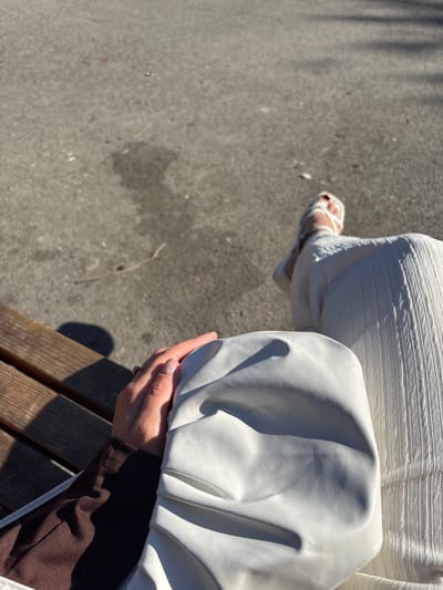 An overhead, medium shot shows a person's legs and hand on a park bench in Belovo, Russia. The ground is paved with grey asphalt, with some debris and shadows from trees. A person's right leg, clad in white ribbed pants and white sandals, extends into the frame. Their left arm, wearing a dark brown long-sleeved shirt, rests on a white clutch bag. The person's hand, with light pink nail polish and a ring on the index finger, is seen holding the bag. The bench is made of wooden planks.