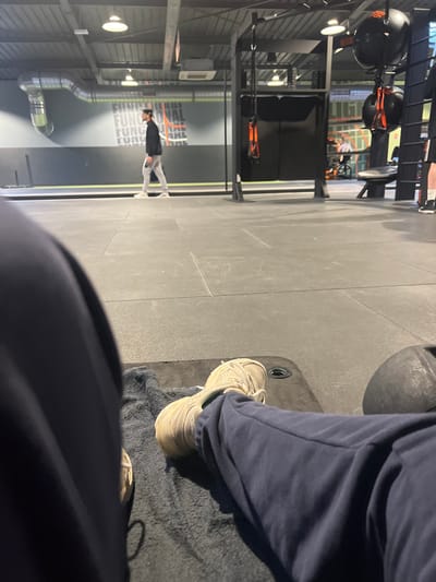 The scene depicts an interior view of a fitness facility located in Annœullin, France. The perspective is from a low angle, showing the lower body of an individual resting on a dark grey towel placed on a dark grey rubberized floor. The individual wears dark blue sweatpants and light beige sneakers. A dark, circular object, possibly a weight, is partially visible to the right of the feet.

In the midground, a male subject with dark hair, wearing a dark long-sleeved top, light grey sweatpants, and white sneakers, walks from left to right across the open gym floor. Behind him is a dark grey wall featuring a repeating horizontal graphic of "FUNCTIONAL" in white and orange lettering. The ceiling displays exposed ductwork, circular recessed lighting fixtures, and HVAC units.

Towards the right background, gym equipment is visible, including a black frame structure with orange suspension straps hanging from it, and dark spherical objects, resembling punching bags or medicine balls, suspended from the ceiling. Further back, a person is seated in an orange and black manual wheelchair near additional gym apparatus. A partial view of another individual's lower legs and feet is visible on the far right edge of the frame. The overall environment is well-lit and appears to be actively in use.