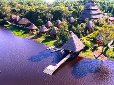 This is an aerial drone shot of a jungle lodge with traditional thatched-roof huts and a larger, tiered pyramid-like structure situated along a wide, dark-water river. Several canoes are moored along the riverbank, near a wooden dock extending into the water. A single person is visible on the dock, and other structures are nestled within the dense, green foliage of the rainforest. The scene is not in Ləcət, Azerbaijan.