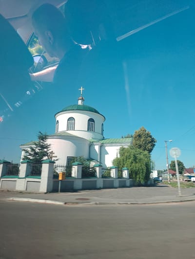 The image shows a white church with a green dome and a cross on top. The church is located in Kotelva, Ukraine. There is a fence in front of the church, and trees and bushes around it. The sky is blue and clear. In the foreground, there is a road and a sidewalk. The image appears to be taken from a moving vehicle, as indicated by the reflection in the window on the left side of the image, which shows the head and shoulder of a person looking out.