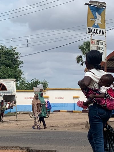 This image captures a street scene in Shendam, Nigeria, under an overcast sky. In the foreground, positioned on the right, a woman with dark hair, wearing a white shirt and blue jeans, walks away from the viewer, looking slightly towards the right. A baby, wearing a white shirt and pink Crocs, is secured to her back with a pink and black patterned fabric carrier, its head resting on the woman's right shoulder. The woman carries a black bag in her left hand.
To the left of the foreground woman, in the midground, two women are walking from left to right on an unpaved surface adjacent to a paved road. The lead woman wears a light brown head covering and a patterned dress, balancing a translucent container on her head and holding an item in her hands. The woman behind her wears a green head covering and a green top, also holding an object. Behind them, a light-colored wall with a blue stripe along its base displays repeating red text that appears to be "NO SMOKING." A bicycle is partially visible behind the second woman.
Further into the background, on the right, a tall roadside sign for "OKACHA" is prominent. The sign features a yellow background with a blue 'X' motif and a white bird or dove-like figure in the center. Below "OKACHA," a smaller sign indicates "PETROL" and "DIESEL," with "840" visible above "PETROL," likely denoting fuel prices. Electrical power lines stretch across the cloudy sky above the street. Green foliage from trees is visible on both sides of the OKACHA sign and in the far-left background. On the far left, a makeshift stall or small shop, appearing to sell various goods, stands by the roadside, with a person's leg partially visible near it. The ground consists of a dark paved road and lighter unpaved sections.