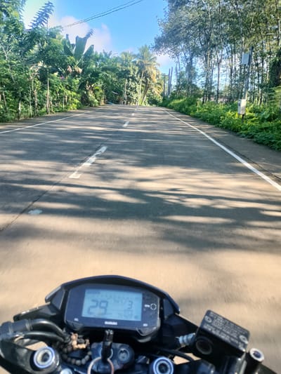 The image captures a first-person perspective from a motorcycle traveling on a paved road lined with lush green foliage and tall trees. The road has white lane markings, and the sunlight casts dappled shadows on the asphalt, indicating a bright day. The sky is blue with scattered white clouds. In the foreground, the motorcycle's dashboard is visible, displaying a digital readout with numbers like "29" and "74/3". The location is identified as Kidapawan City, Philippines, suggesting a tropical or subtropical environment. The overall scene depicts a journey through a scenic, tree-lined road.