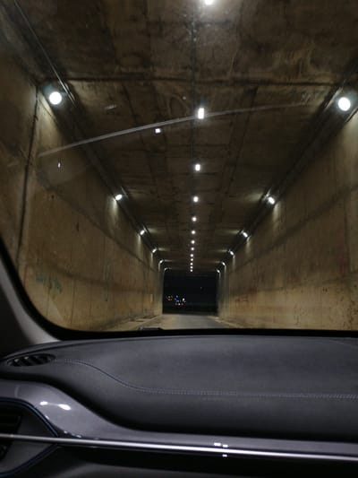 The image shows the view from inside a car as it drives through a concrete tunnel. The tunnel is illuminated by rows of lights on the ceiling and walls. The walls of the tunnel are rough concrete with some graffiti. The interior of the car is visible in the foreground, including the dashboard and windshield. The time of day appears to be nighttime, as the tunnel exit in the distance is dark with some distant lights. The weather is not discernible. The location is Purbachal, Bangladesh, as indicated by the provided context. No people are visible in the image.