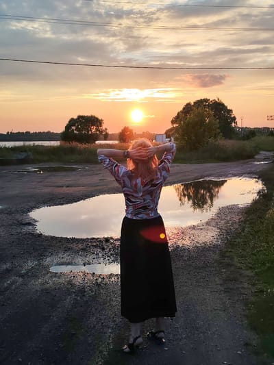 The image depicts a woman standing with her back to the camera, facing a sunset over a body of water. She has her hands behind her head, and is wearing a patterned blouse and a long black skirt. She is standing on a dirt path that has several puddles reflecting the sky. In the background, there are trees and a body of water. The sky is filled with clouds and the sun is low on the horizon, casting a warm orange glow. The location is the city of Chelganovo, Russia.