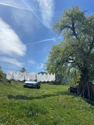 The image depicts a rural scene, likely in Đurđevića Tara, Montenegro. A white car, obscured by white sheets hanging on a clothesline, sits in a field of grass dotted with white and yellow wildflowers. Behind the car and clothesline rises a tree with lush green foliage. The background reveals partially visible mountains and a pile of wood. The sky is blue with contrails.
