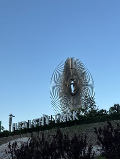 A low-angle view showcases a metal sculpture resembling an abstract eye against a clear blue sky. The sculpture is composed of concentric metal rings and radiating metal lines. To the right of the sculpture, the name "FAITH HAYARD PARK" is displayed vertically in large, white capital letters, bordering a brick wall covered in greenery and shrubbery. Elements suggesting park infrastructure are visible below the park's name. The location is Taihe District, China.
