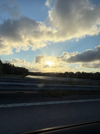 The image depicts a solar panel farm situated in Ödsmål, Sweden, during sunset. The foreground shows a highway barrier, indicating the viewer is likely traveling by car. Beyond the barrier, rows of solar panels are visible, stretching across a field backed by a dense line of trees. The sky is filled with dramatic clouds illuminated by the setting sun, casting a warm glow over the landscape. The overall scene suggests a rural or semi-rural environment in Sweden.