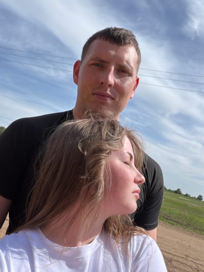 A man and a woman are lying on the ground in a field. The man is wearing a black t-shirt, and the woman is wearing a white t-shirt. The woman is lying on her back with her eyes closed. The man is looking at the camera. The sky is cloudy. There are power lines overhead. The location is near the city of Ural'skoe, Russia. The scene is a close-up shot. The ground appears to be dirt and grass.