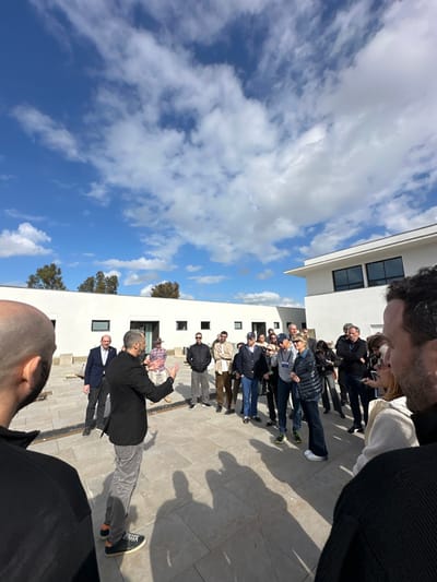 An outdoor scene in Kadima-Zoran, Israel, depicts a group of approximately 20-25 individuals gathered on a light-colored paved courtyard under a bright blue sky with scattered white clouds. A male speaker, wearing a dark blazer and grey trousers, stands centrally, facing the group and gesturing with both hands. The audience, composed of men and women of varying ages dressed in casual to smart-casual attire, forms a semicircle around him, observing the speaker. Some individuals wear lanyards or sunglasses. Two partial figures are visible in the immediate foreground, positioned behind the camera's perspective, observing the scene. In the background, two white, modern, flat-roofed buildings are visible: a long, single-story structure with multiple small rectangular windows on the left and a two-story building with larger dark-framed windows on the upper floor on the right. Trees are present in the far left background. Long shadows of the people are cast on the pavement, indicating strong sunlight from a low angle. The overall environment suggests a contemporary architectural setting.