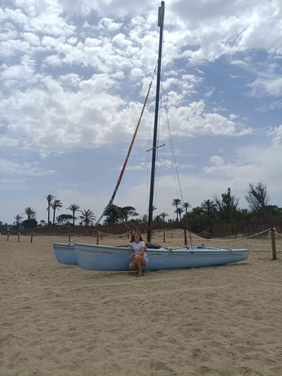 A young woman with long brown hair, wearing a white t-shirt and brown shorts, is crouching on a sandy beach next to a blue catamaran. The catamaran is beached on the sand, and its tall mast is raised, with a red, white, and blue striped sail attached. The woman is looking at the camera, with her right hand resting on the side of the boat. Behind the catamaran, palm trees and other vegetation line the shore. The sky is partly cloudy with a mix of blue sky and white clouds. Wooden posts with rope are visible in the foreground, demarcating a pathway on the beach. The scene appears to be in Sagunto, Spain, a coastal city known for its beaches and Roman history.