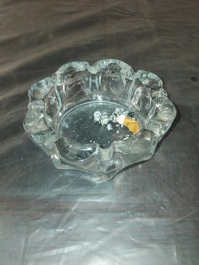 A glass ashtray with a floral rim is situated on a dark, possibly plastic, surface. The ashtray contains a partially smoked cigarette and ash. The surface appears to be a dark, textured material with some reflective qualities. The lighting is dim, creating shadows and highlighting the texture of the surface and the ash within the ashtray. The image is taken from a slightly elevated angle, looking down into the ashtray. The location is specified as Chhina Uttar, Pakistan, but this detail is not visually evident from the image itself.