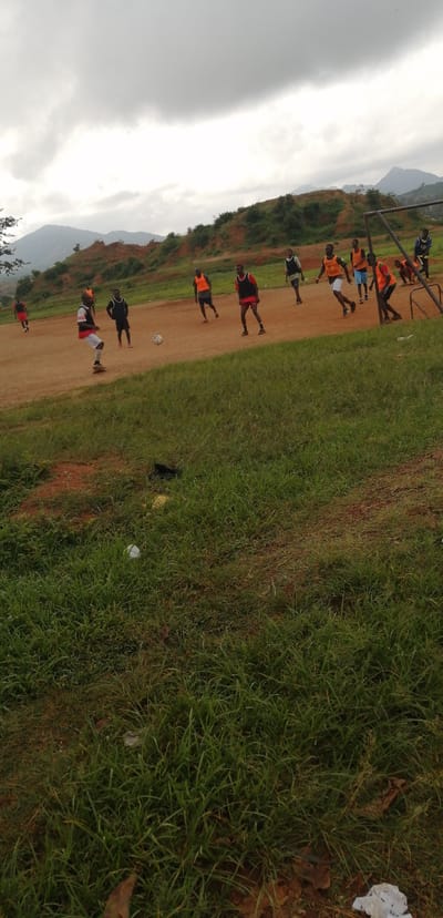 A group of young men are playing soccer on a dirt field. The field is surrounded by grassy areas and hills in the background. The sky is overcast. One player is in the foreground kicking the ball. Another player is to his right, wearing an orange bib. Several other players in orange and black bibs are scattered across the field, engaged in the game. A soccer goal is visible on the right side of the image. The overall setting appears to be a rural or semi-rural area in Barakin, Nigeria.