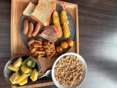 A wooden tray displays a variety of breakfast foods. A white bowl contains cereal with milk. A dark bowl holds pineapple chunks and kiwi slices. A grey plate features two fried shrimp, slices of ham, three sausages, two pieces of toast, small fried balls, and two pastries. A silver spoon rests on the tray next to the cereal.