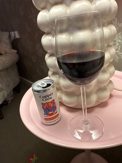 The image captures an indoor setting within a residence in Babergh, United Kingdom. The primary focus is a wine glass containing dark red wine, approximately half full, positioned on a round, light pink side table or tray. Adjacent to the wine glass is an opened aluminum can, labeled "The Copper Crew Negroamaro Organic Red Wine," also resting on the pink surface. A prominent white, multi-lobed ceramic lamp or decorative object occupies the background behind the glass and can. To the far left, a portion of a light-colored upholstered armchair is visible against a patterned brown wall. A white electrical cable extends from behind the lamp across the pink surface. On the dark carpet in the lower-left foreground, a small red cherry-shaped decorative object is present. No individuals are depicted in the scene.