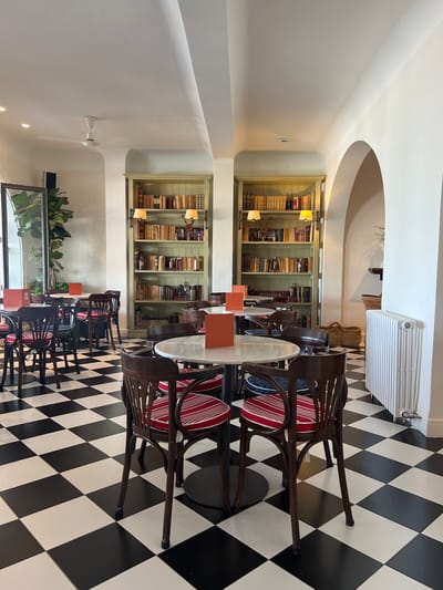 This image depicts the interior of a cafe in Port de Pollença, Spain. The cafe features a striking black and white checkered tile floor. Several round marble-topped tables are arranged throughout the space, each surrounded by wooden chairs with red and white striped seat cushions. Two tall, pale green bookshelves filled with books are prominently displayed against a white wall. One bookshelf has two lamps attached to it. In the background, an arched doorway leads to another area of the cafe, and a white radiator is visible on the wall. A large plant in a pot is situated near a glass door on the left side of the image. The lighting is bright, suggesting daytime. There are no people visible in the scene.