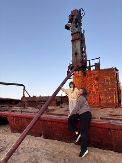 Two young females are posed on the deck of a large, rusted ship resting on a sandy beach under a clear blue sky. The ship appears to be a derelict vessel, with extensive rust and decay evident on its structure. One female, wearing a yellow jacket and grey pants, stands near a prominent metal pipe, her left hand resting on it. The other female, wearing a grey hoodie and black pants, is seated on the edge of the ship's deck, her legs dangling towards the sand. The scene is set in Teriberka, Russia, known for its stark coastal landscapes and ship graveyards. The overall environment suggests a remote, possibly abandoned location.