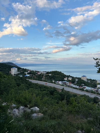 The image depicts a picturesque coastal landscape with a town nestled on a hillside overlooking a vast body of water. The sky is a canvas of blues and whites, with scattered clouds catching the soft light of either dawn or dusk, suggesting a serene time of day. The foreground is dominated by lush green foliage and rocky outcrops, hinting at a natural, untamed environment.

A winding road snakes its way along the hillside, bordered by streetlights and a white barrier, suggesting a well-developed, albeit scenic, thoroughfare. Along this road, various buildings are visible, ranging from smaller houses with colorful roofs to a taller, modern apartment building. These structures blend into the hillside, suggesting a harmonious integration of human habitation with the natural surroundings.

The expansive body of water in the background could be the Black Sea, given the location context of Malyi Mayak, Ukraine. The water appears calm and stretches towards a distant horizon, where it meets a hazy sky. The overall impression is one of tranquility and natural beauty, with a touch of human presence. There are no visible people or specific activities taking place, the scene is static, focusing on the landscape and architecture. The weather appears clear and pleasant, with the soft lighting contributing to a peaceful atmosphere.