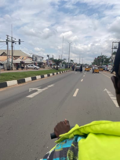 The image depicts a street scene in Karu, Nigeria. In the foreground, a person's hand is visible, holding onto a handlebar, suggesting the viewpoint is from a vehicle, possibly a motorcycle or a tricycle. The person is wearing a bright yellow vest over a patterned garment.
The road is paved and has lane markings. On the left side of the road, there are buildings with shops, including one with a sign that reads "VIP Deluxe Lounge." There are streetlights, utility poles with wires, and a traffic light visible. Some pedestrians are walking along the sidewalk and crossing the road. A silver car is parked near the shops.
On the right side of the road, several yellow tricycles, commonly known as "Keke Napep," are present, some with passengers. More pedestrians and shops are visible further down the street. The sky is cloudy. The overall environment suggests a bustling urban or semi-urban area.