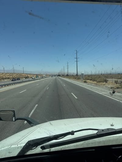 The image is a point-of-view shot from the cab of a truck driving on a highway, likely in Palm Springs, California, United States, given the presence of wind turbines and the arid landscape. The sky is clear blue. A white truck hood and windshield wipers are visible in the foreground, indicating the perspective of the driver. The highway stretches into the distance, with multiple lanes of traffic, including other cars and trucks. On either side of the highway, there is dry, scrubby vegetation and hills. A prominent feature of the landscape is a large number of wind turbines, characteristic of the area around Palm Springs. Power lines on tall poles run parallel to the highway. A speed limit sign indicating "55" is visible on the right side of the road. The overall scene depicts a typical highway drive through a desert region with renewable energy infrastructure. There are several specks and smudges on the windshield, obscuring the view slightly.