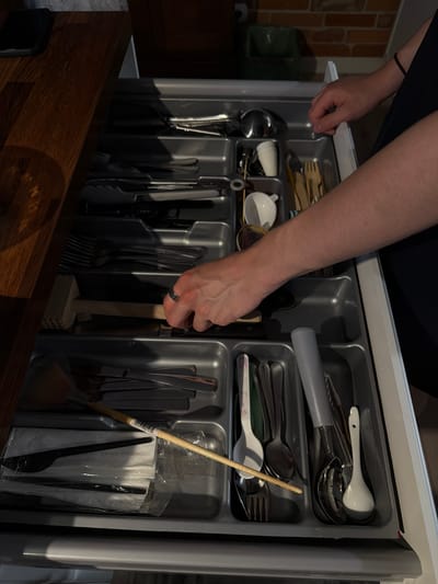 A person's hands are shown reaching into an open kitchen drawer filled with cutlery. The drawer organizer is made of grey plastic and is divided into compartments. Various utensils, including spoons, forks, knives, and what appears to be a meat tenderizer with a wooden handle, are neatly arranged within the compartments. Some cutlery is stacked, while others are placed individually. A white ceramic cup and a white spoon are visible in one compartment. The background shows a brick wall and a dark wooden surface. The lighting suggests it is daytime, with natural light casting shadows across the drawer. The scene is set in a kitchen. No visible text is present in the image.