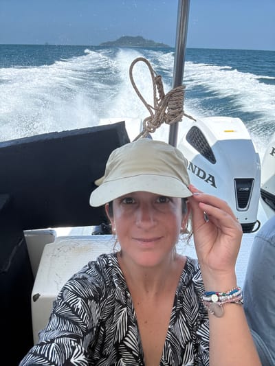 The scene depicts a daytime marine environment from the perspective of a moving boat, identified as near Pak Nam, Thailand. A female subject occupies the central foreground, looking directly at the viewer with a slight smile. She wears a light beige sun hat, a black and white patterned top with a deep V-neck, and multiple beaded bracelets with a silver charm on her left wrist. Her left hand is raised, touching the brim of her hat. To the right, a partial view of another individual's arm, wearing a light blue garment, is visible. In the background, two white Honda 250 horsepower outboard motors are mounted at the boat's stern, generating a substantial white wake across the blue ocean surface. A thick, light-colored rope is coiled and secured to a vertical metal stanchion. On the distant horizon, a small, tree-covered island is present under a clear sky.