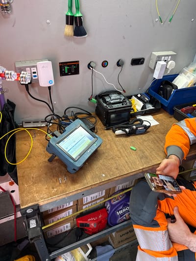 The image shows the interior of what appears to be a work van, with an individual in high-visibility orange and grey workwear on a video call on their smartphone. The van contains various tools and equipment for fiber optic work. A fiber optic fusion splicer and an EXFO MaxTester are prominent on a wooden surface. The van also contains a distribution box, network accessories from TP-Link, brushes, and several boxes, including a Cadbury HERO selection. The environment is cluttered with cables and tools. No specific location details relating to Mid Suffolk, United Kingdom, are visible.