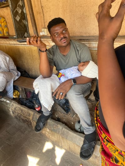 In Sabon Gari South, Nigeria, an adult male is seated outdoors or in an open-fronted structure, holding an infant. The man has a dark complexion, short dark hair, a mustache, and a goatee. He wears a dark olive green t-shirt, light grey trousers, black socks, and black sandals. A black beaded bracelet is on his left wrist, a smartwatch with a teal band on his right wrist, and a dark-stoned ring on his right ring finger. He holds a black remote control in his left hand. His facial expression indicates a focused or contemplative gaze upwards and to his right.
The infant, held in the man's right arm, is dressed in a light blue and white patterned garment and a white knitted cap, appearing to be asleep. A small, colorful toy with multiple segments is visible near the infant's chest.
To the man's immediate left, another individual is partially visible, seated, wearing light-colored clothing. To the man's right, the arm and shoulder of a third person are visible in the foreground, wearing vibrant red, yellow, and black patterned fabric. This person's right arm is raised, suggesting an active gesture or interaction.
The subjects are seated on a wooden bench. Behind them is a textured, light brown wall. Below the bench, a concrete ledge borders a dirty area containing black plastic bags, what appears to be a partially filled red Coca-Cola bottle, and other debris. To the far left, stacked yellow packaged goods are visible on a shelf, along with a dark patterned fabric. A white electrical outlet is mounted on the wall behind the man. The ground in the foreground is paved with light grey blocks, with patches of sunlight illuminating the surface.