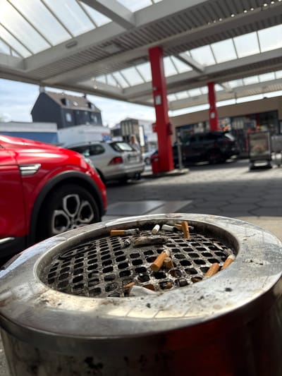 The image shows a close-up of a metal ashtray filled with cigarette butts. Behind it, slightly blurred, are a red car and other vehicles. The background suggests an indoor car repair or service center, with red support beams and a glass ceiling structure. The scene is likely located in Wuppertal, Germany. There are no people visible.