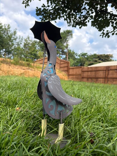 A dark gray metal garden sculpture resembling a duck stands in a patch of green grass. The duck is facing to the right, with its head tilted upwards and a black umbrella held over it. The umbrella has a spiky edge, and the duck's beak is a reddish-brown. The duck has a teal blue panel with a swirling pattern on its body, and its wings are detailed with gray lines. It stands on two yellow boots. In the background, there is a brown wooden fence, trees, and a cloudy blue sky. The scene is set in Stallings, North Carolina, United States of America.