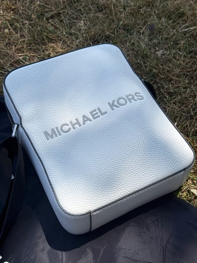 A white, textured leather crossbody bag with "MICHAEL KORS" embossed in a silver-grey font is lying on dry grass. The bag is angled, with the top left corner closer to the viewer. A black strap is attached to the bag and disappears into the lower right corner of the frame. A small zipper pull with the "MICHAEL KORS" logo is visible along the bottom edge of the bag. Next to the bag on the right is a dark, possibly black, fabric surface. The scene appears to be outdoors in natural light, suggesting it's daytime. The dry grass and natural lighting might indicate a warm season or a location with dry vegetation. The overall impression is a casual setting.