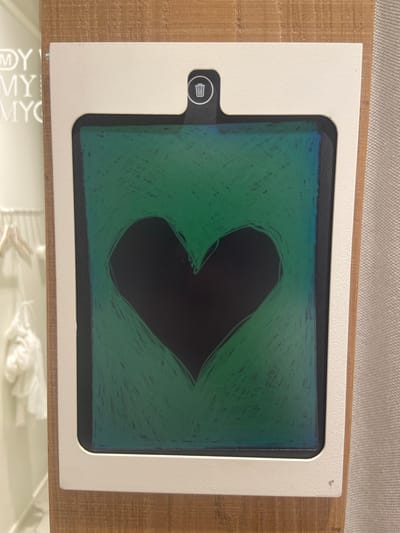 The image shows a white digital drawing tablet with a black frame, set on a wooden surface. The screen displays a drawing of a heart in dark green with blue accents at the top. The heart shape is outlined and filled with scribbled lines, suggesting a child's artwork. To the left of the screen, there is a small, white icon resembling a trash can within a black circle, indicating a delete function. The tablet is positioned on a light brown wooden table, and behind it, a textured grey surface is visible, possibly a wall or fabric. Below the tablet, a white surface with repeated blurry text "MYC" is visible. The overall setting appears to be indoors, likely a home or a child's play area. No specific time of day or weather conditions can be determined. The emotion conveyed is playful and creative, typical of a child's drawing. The visible text is "MYC" which is blurry and repeated.