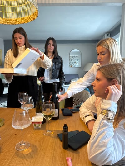 The image depicts four adult females engaged in a social gathering within a residential interior, specifically identified as being in the city of Saint-Étienne, France. The exact address within Saint-Étienne is not discernible.
In the foreground, a wooden table occupies the central space, holding several objects. These include three clear stemmed wine glasses, one containing white wine, and two dark-colored wine bottles, one appearing full and the other partially consumed. Also present on the table are a small white rectangular container labeled "CHAVROUX," two dark-colored smartphones, a black vape device, and a pink lip gloss tube. A small green bowl is partially visible on the far left of the table.
Four females are present. On the far left, a female with dark brown hair, wearing a white ribbed turtleneck and a black skirt, is in the process of opening a white rectangular box. Next to her, a female with dark hair, dressed in a black leather jacket over a white shirt, stands observing the box. To the right of the scene, another female with blonde hair, wearing a white button-up shirt, leans over the table, reaching towards the wine bottles. In the immediate foreground, facing right in profile, a blonde female wearing a white button-up shirt with prominent cuffs and multiple bracelets, including a large gold-toned watch, is seated. Her right hand is raised to her ear, and she appears to be listening or deep in thought, looking away from the central interaction.
The background shows a brightly lit room with light-colored walls, one featuring a bluish-grey accent. A large, woven, light-colored pendant lamp hangs from the ceiling. An arched mirror on the back wall reflects a patterned wallpaper. A blue sofa or piece of furniture is partially visible in the distant background. The overall environment suggests a casual gathering in a contemporary living or dining area.