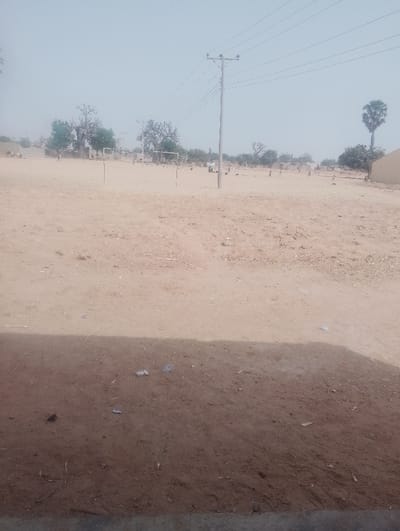 The scene depicts an expansive, dry dirt field in Kanwa, Nigeria, under a hazy, pale blue sky. The immediate foreground consists of darker brown soil with scattered small refuse. The larger midground field is a lighter brown, featuring two white metal football goalposts positioned in the center-left. Numerous small, indistinct human figures are scattered across this field, some in clusters, others individually. A tall, grey utility pole, likely concrete, with multiple overhead power lines, stands in the mid-right, with lines extending horizontally across the upper frame. The background reveals sparse trees, some with visible green foliage and one tall, slender palm tree on the far right. Several single-story, light-colored buildings are interspersed among the distant trees.