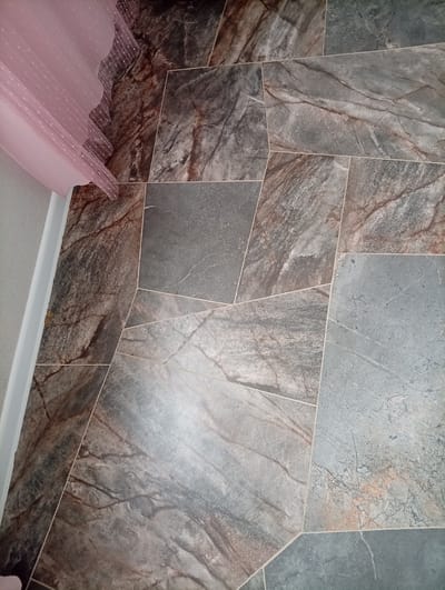 The image shows a close-up view of a floor covered with large, irregularly shaped tiles that resemble natural stone with a marbled pattern in shades of gray, brown, and blue. The tiles are laid in a somewhat random pattern, with thin grout lines between them. In the upper left corner, a pink, sheer fabric with a dotted pattern is draped over the edge of the floor, partially obscuring the wall. The fabric has a scalloped edge. The lighting suggests an indoor setting, possibly a room with natural light. There is no discernible activity or event taking place. The context provided suggests the location is Çolpon-Ata, Kyrgyzstan.