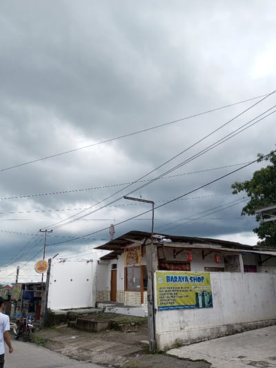 This image captures a street scene in Cibadak, Indonesia, under a heavily overcast sky. In the lower left foreground, the back of an individual wearing a white t-shirt is visible as they walk on a paved surface. Adjacent to their path, a red and black motorcycle is parked.

The midground features a series of commercial structures and infrastructure elements. A prominent single-story building, likely a shop, has a corrugated metal roof and a light-colored facade. A large yellow banner reading "BARAVA SHOP" in blue text and displaying images of apparel, including jeans, is affixed to a concrete wall in front of this building. Several red decorative lanterns hang near the building's roofline. To the left, another white-painted structure with a brown wooden door and a yellow circular sign is visible, though its text is unreadable.

Numerous black electrical and communication wires crisscross the upper and middle portions of the frame, suspended from utility poles. Several streetlights are also present. A cellular tower is partially visible in the background, behind the buildings. On the right, green foliage from a tree extends above a concrete wall. The ground is a mix of uneven paved sections and bare earth with minimal vegetation. Faint outlines of distant hills are discernible under the cloudy horizon.