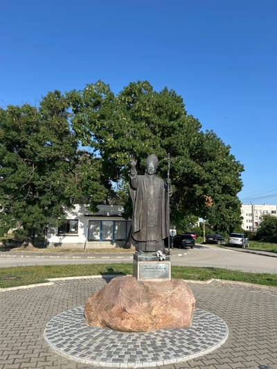 This media file features a bronze statue of a religious figure, likely a pope, holding a staff. The statue is positioned outdoors, in a paved area that incorporates a large, reddish-brown boulder. A metal plaque with visible text is attached to a pedestal near the boulder. The statue and its surroundings are bathed in sunlight, suggesting it is daytime. Behind the statue, lush green trees are prominent, with a building and several parked cars visible in the background. The sky is a clear, bright blue, indicating fair weather. The overall scene conveys a sense of public art installation within an urban or suburban environment. The visible text on the plaque reads "Joannes Paulus II. Chosman Missae Theod. 1920 - 2005 - Benedictus XVI". This indicates the statue is dedicated to Pope John Paul II, and mentions Pope Benedict XVI, suggesting a memorial or tribute. The location context provided is Belene, Bulgaria.