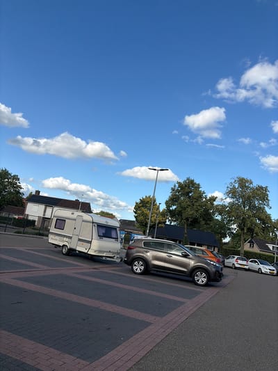 The image depicts a parking lot in Eibergen, Netherlands, on a clear day with scattered white clouds against a bright blue sky. In the foreground, a gray SUV is parked to the right, angled towards the viewer. To the left of the SUV, a white caravan is hitched to an unseen vehicle, its front facing away from the viewer and slightly angled. The caravan is positioned on a dark asphalt surface with red brick borderings forming parking spaces.
In the background, several houses with dark tiled roofs are visible, some with solar panels on the roof. Lush green trees are interspersed between the houses, and a tall lamppost stands in the middle ground. A few other cars are parked in the background, including a white hatchback and an orange car. The overall scene suggests a typical suburban or commercial area in Eibergen. There are no people visible in the image.