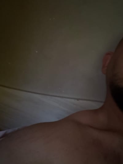 The image shows a close-up, dimly lit view of a person's shoulder and part of their head. The background is a textured, light-colored surface, possibly a ceiling or wall, with some faint dust specks visible. A dark, horizontal line runs across the mid-ground of the image. The person appears to be lying down or reclining, with their shoulder exposed. The person has dark hair and a beard. No specific location in Borovan, Bulgaria, can be identified from the image.