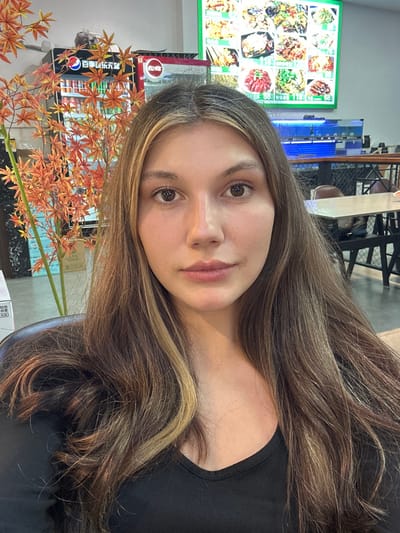 A female subject is centered in a close-up indoor photograph taken within a casual dining establishment in Huangdao District, China. The subject, a young adult with light skin, brown eyes, and long, multi-toned brown hair with blonde highlights, faces forward with a neutral expression, wearing a black v-neck top. To her left, an artificial orange-red maple-leaf plant is positioned in front of a glass-fronted commercial beverage refrigerator, branded with Chinese text ("百事可乐" - Pepsi Cola) and stocked with bottled and canned drinks. A second red-framed commercial cooler is partially visible adjacent to the first. To her right, an illuminated menu board displays various food dishes with images and Chinese text against a green background. Below the menu board, a blue-lit aquatic display tank is visible. The far right background reveals empty dining tables with dark surfaces and dark chairs, consistent with an unoccupied restaurant seating area.