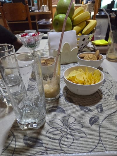The image displays an indoor tabletop scene located in Negorci, North Macedonia. The surface is covered by a light-colored placemat adorned with a repeating gray floral pattern. In the left foreground, two empty, tall, clear, ribbed drinking glasses are visible. To their right, a third clear glass contains a small volume of frothy, light-brown liquid and a pink-and-white striped drinking straw. Further right, two small white bowls are present; one contains yellow potato chips, and the other holds light-brown, round cookies. A white, perforated metal bowl in the mid-background is filled with green apples and yellow bananas. Adjacent to this, a white, flower-shaped napkin holder contains folded white napkins. A footed crystal bowl filled with red and white decorative elements is partially visible behind the foreground glasses. On the far right, a tall, mostly empty clear glass with a straw stands next to a black-and-clear shaker bottle. A yellow sticky note is partially visible beneath the shaker bottle. A dark-colored sleeve or arm is partially visible on the far left edge of the frame, indicating a person's presence. The scene is illuminated by artificial indoor lighting.