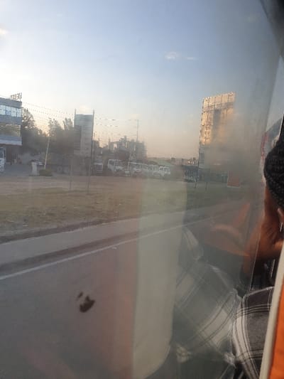 A blurry, upward-angled shot depicts a roadside scene through a vehicle window. Buildings of varying heights line the road, including a notable billboard. Vehicles are parked along the side of the street. The sky is light, suggesting it is either early morning or late afternoon. The window's surface has a few smudges. The upper portion of the image captures part of the vehicle's interior, specifically a person dressed in casual wear.