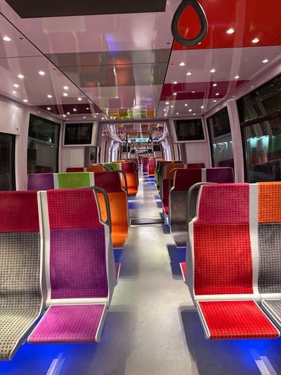 The image shows the interior of a train, likely a suburban train, in Saint-Brice-sous-Forêt, France. The train car features rows of seats upholstered in a variety of bright colors, including red, orange, green, and purple, each paired with gray fabric on the lower section of the seatback. The seats are arranged facing each other along the length of the car.
The walls of the train car are white with vertical red and gray bands, and are punctuated by evenly spaced circular lights. There are also square windows, some of which have integrated screens. The floor is a light gray color. The angle of the photo creates a sense of perspective, with the rows of seats converging in the distance.