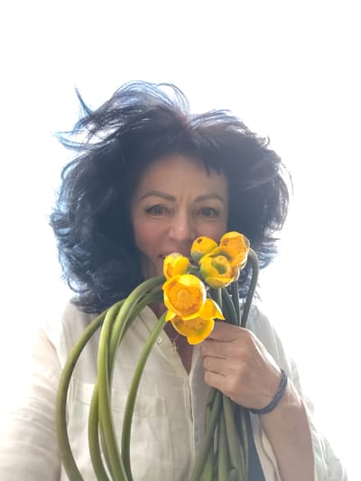 In this medium shot, a woman with dark, voluminous hair styled with a dramatic flip looks directly at the viewer. She is holding a bouquet of yellow flowers with thick green stems close to her face, partially obscuring her mouth. Her expression is playful and engaging. She is wearing a light-colored, button-up shirt. A dark bracelet is visible on her right wrist. The background is brightly lit, suggesting it might be daytime with strong natural light, possibly outdoors or near a window. The overall mood is cheerful and natural. The location context is Мётчанскі сельскі Савет, Belarus.