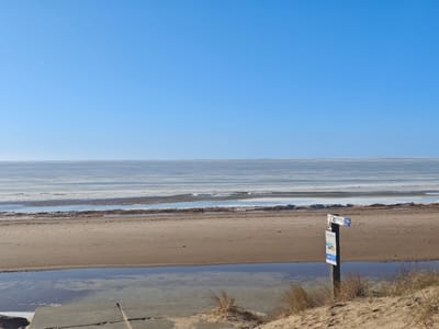 The scene depicts a frozen or partially frozen coastline in Vitrupe, Latvia, under a clear, bright blue sky. The upper half of the image is dominated by the unobstructed sky, with a faint white contrail visible in the upper left quadrant. The horizon is flat and distinct, separating the sky from the expansive body of water.

The water extends from the distant horizon to the sandy beach, largely covered by sheets of light gray and white ice or frozen slush, interspersed with darker patches of open water or thin ice. The shoreline consists of a wide, light brown sandy beach. In the mid-ground, a stretch of standing water or a shallow channel runs parallel to the main body of water on the beach itself.

In the immediate foreground, on the left, a section of a concrete or paved ramp is visible, descending towards the standing water on the beach. On the right, a sandy dune-like rise, covered with sparse, dry, light brown grass, occupies the foreground. Embedded in this sandy rise is a dark wooden signpost. The post features at least three signs: a small, blue rectangular sign pointing right at the top (partially obscured), a larger rectangular informational sign below it with visible text "VITRUPE" and a blue logo/symbol, and another small, blue rectangular sign pointing left at the bottom. No individuals are present in the image.
