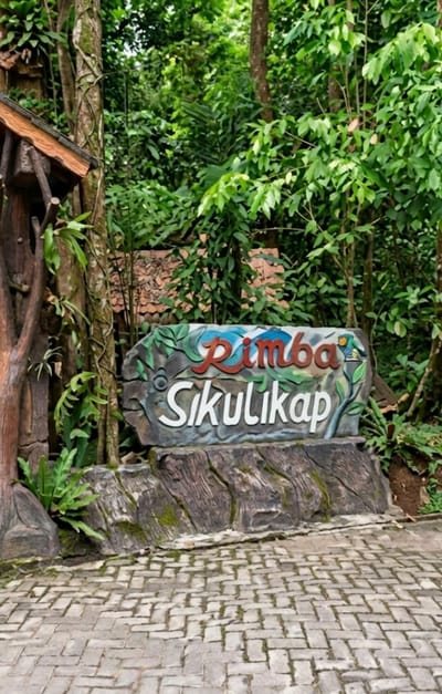 The image shows a sign that reads "Rimba Sikulikap" in a natural environment. The sign is carved into a stone-like material and is decorated with colorful images of what appear to be birds and abstract shapes. The sign is placed on a rocky base, with lush green foliage and trees surrounding it. To the left of the sign, there is a rustic wooden structure, possibly an entrance or a part of a building, with a brown roof and wooden posts. The ground in front of the sign is paved with interlocking bricks, arranged in a pattern. The overall scene suggests an entrance to a natural park, a jungle-themed attraction, or a place named "Rimba Sikulikap" located in the city of Dolat Rayat, Indonesia, which is known for its natural beauty and attractions. There are no people visible in the image. The lighting suggests it is daytime, possibly overcast or in a shaded area due to the dense foliage.